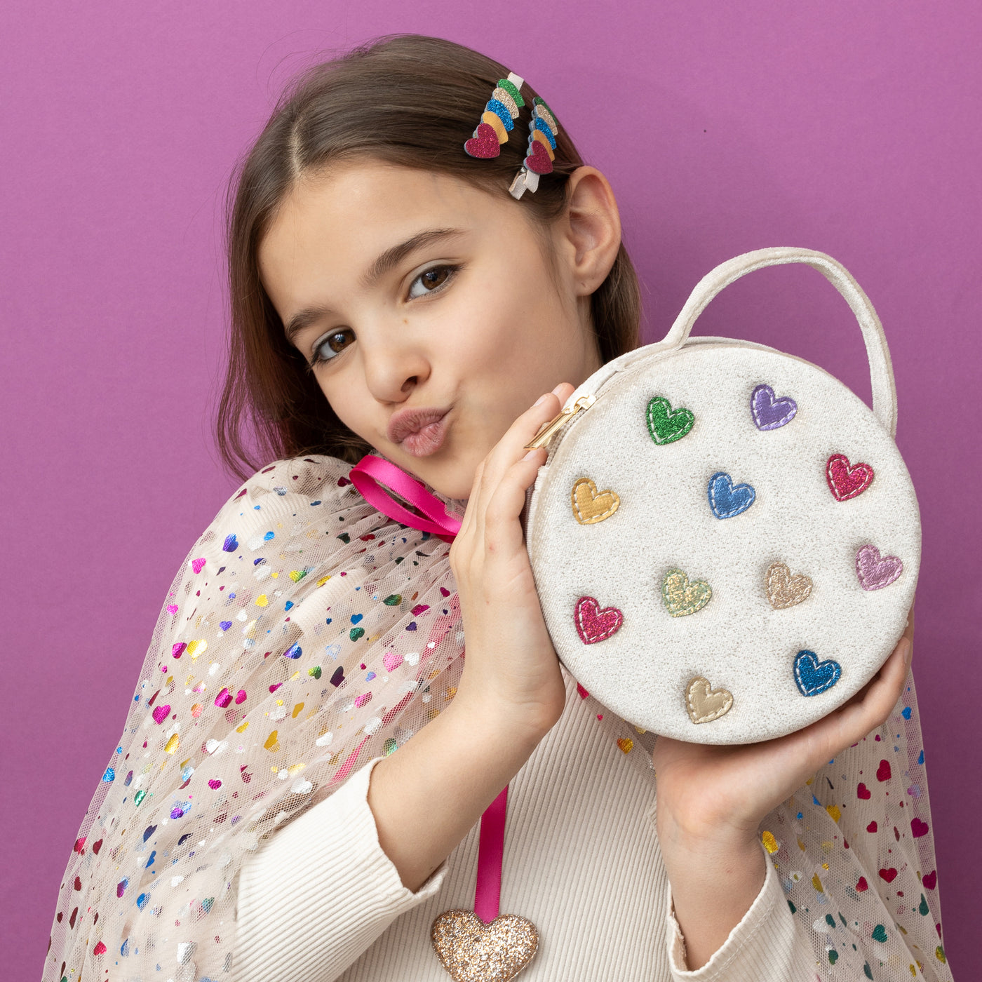 Dark haired girl on purple background holding a cream velvet jazzy heart bag- multi coloured hearts- zip closure- featuring a grab handle and a cross body strap, wearing a rainbow hearts cape and glitter heart hair clips