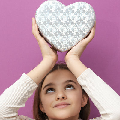 Dark haired girl holding a silver holographic heart bag with zip closure- rope strap with blue glittery heart and pink heart detailing above her head on purple background