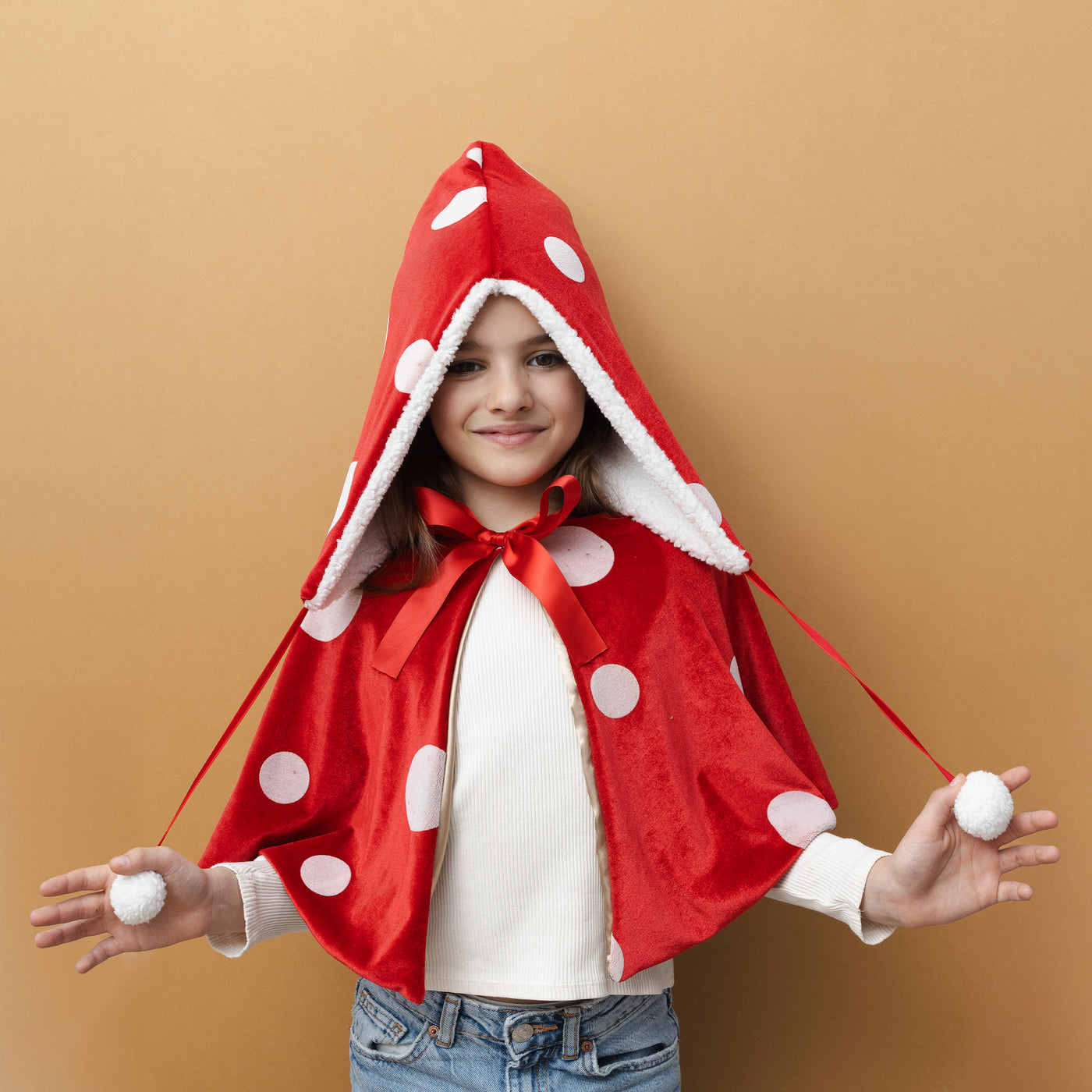 Little girl in jeans wearing a toadstool dress up set featuring red sateen hat with white spots, fluffy borg lining, borg pompoms-Red velvet cape with white spots- gold lining- Red sateen ribbon closure