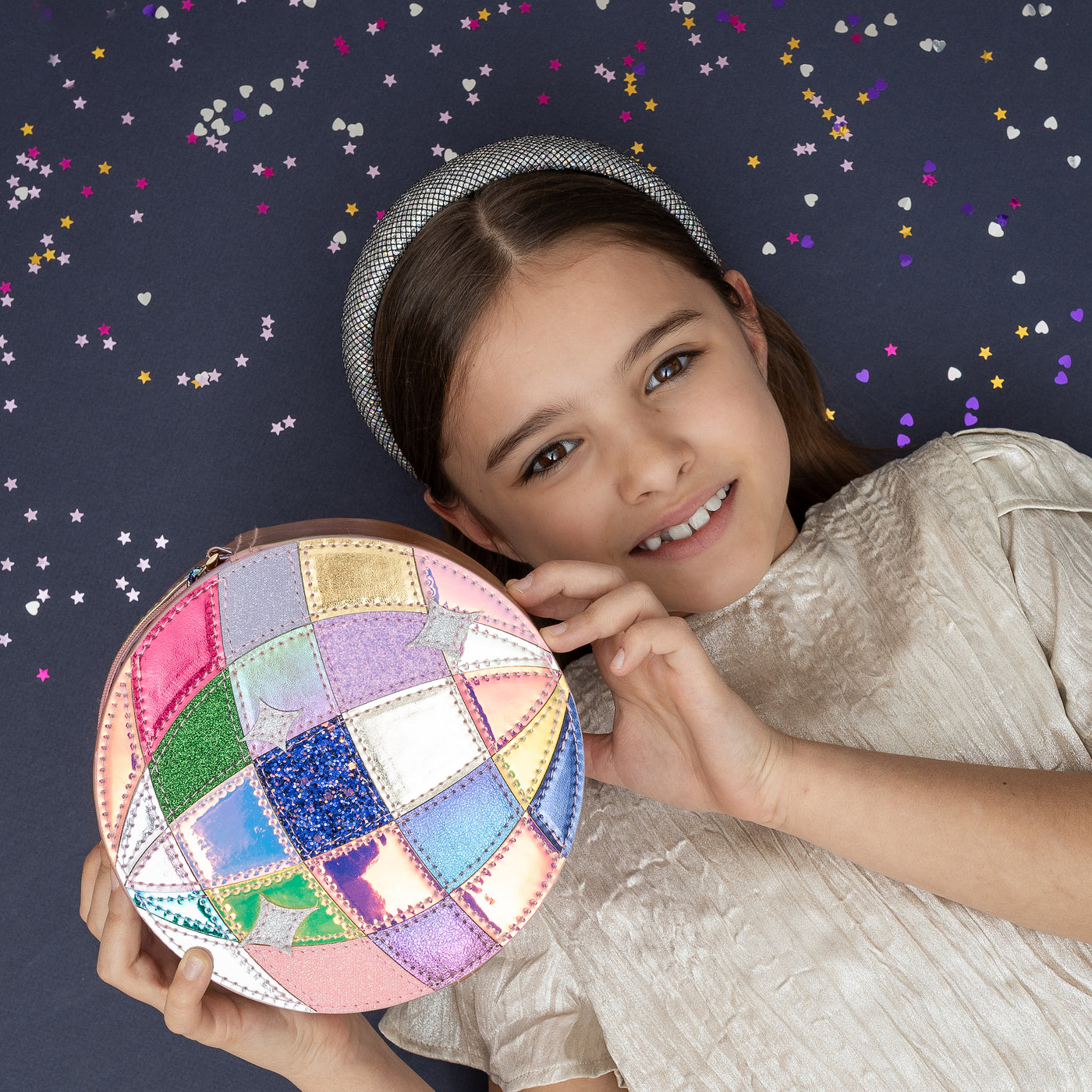 Dark haired girl laid on navy background holding a shimmering pink round disco ball bag- holographic, glitter and PU panels- zip closure- cord strap and a disco theme silver padded alice band