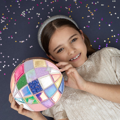 Dark haired girl laid on navy background holding a shimmering pink round disco ball bag- holographic, glitter and PU panels- zip closure- cord strap and a disco theme silver padded alice band
