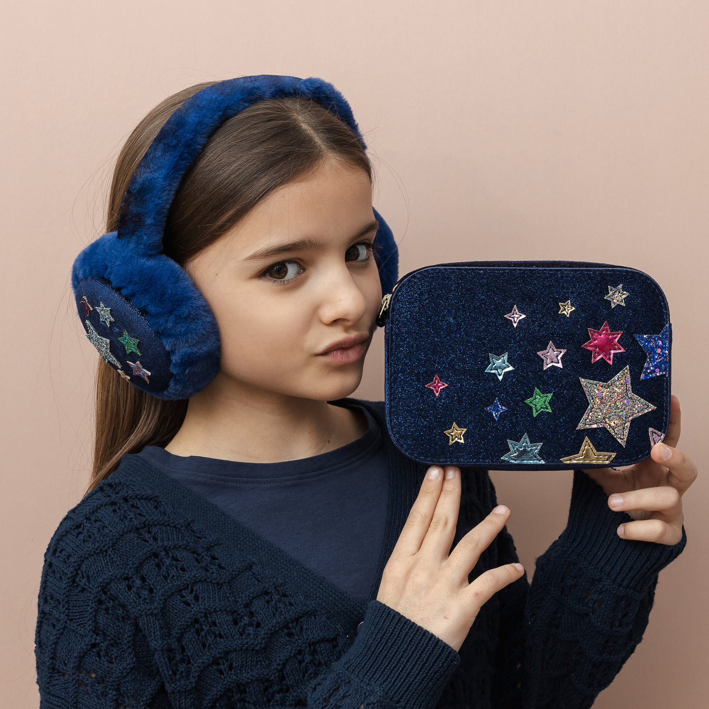 Dark haired girl holding a navy glitter hard shell bag embellished with glitter and metallic multicoloured stars wearing matching navy star embellished earmuffs