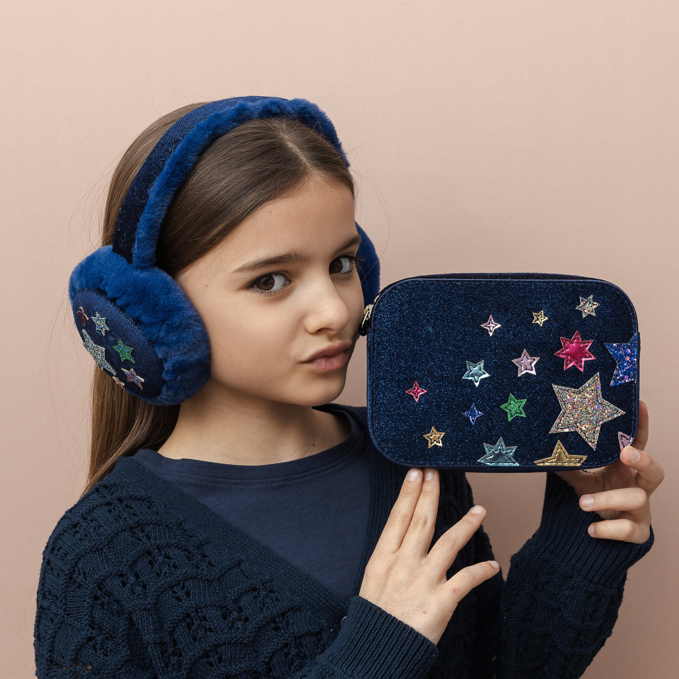 Girl wearing blue earmuffs holding a matching bag with star designs on a beige background