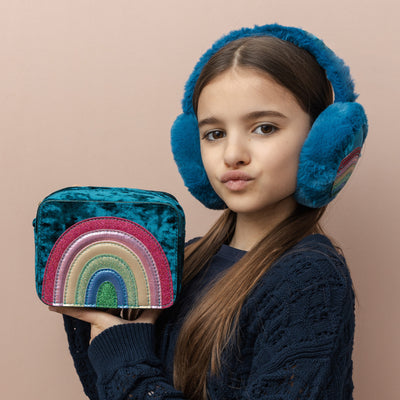 Dark haired girl holding a  Radiant teal rainbow velvet bag with sparkly PU rainbow- zip closure- cross body strap and matching teal fluffy earmuffs with colourful rainbow motif