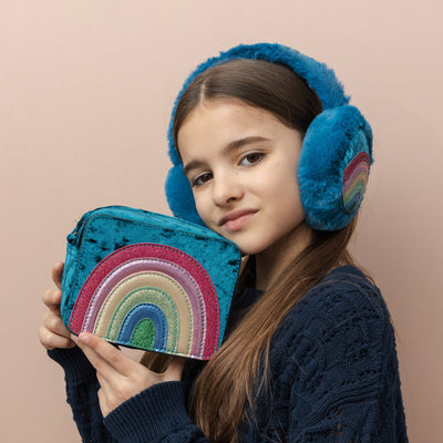 Dark haired girl holding a  Radiant teal rainbow velvet bag with sparkly PU rainbow- zip closure- cross body strap and matching teal fluffy earmuffs with colourful rainbow motif