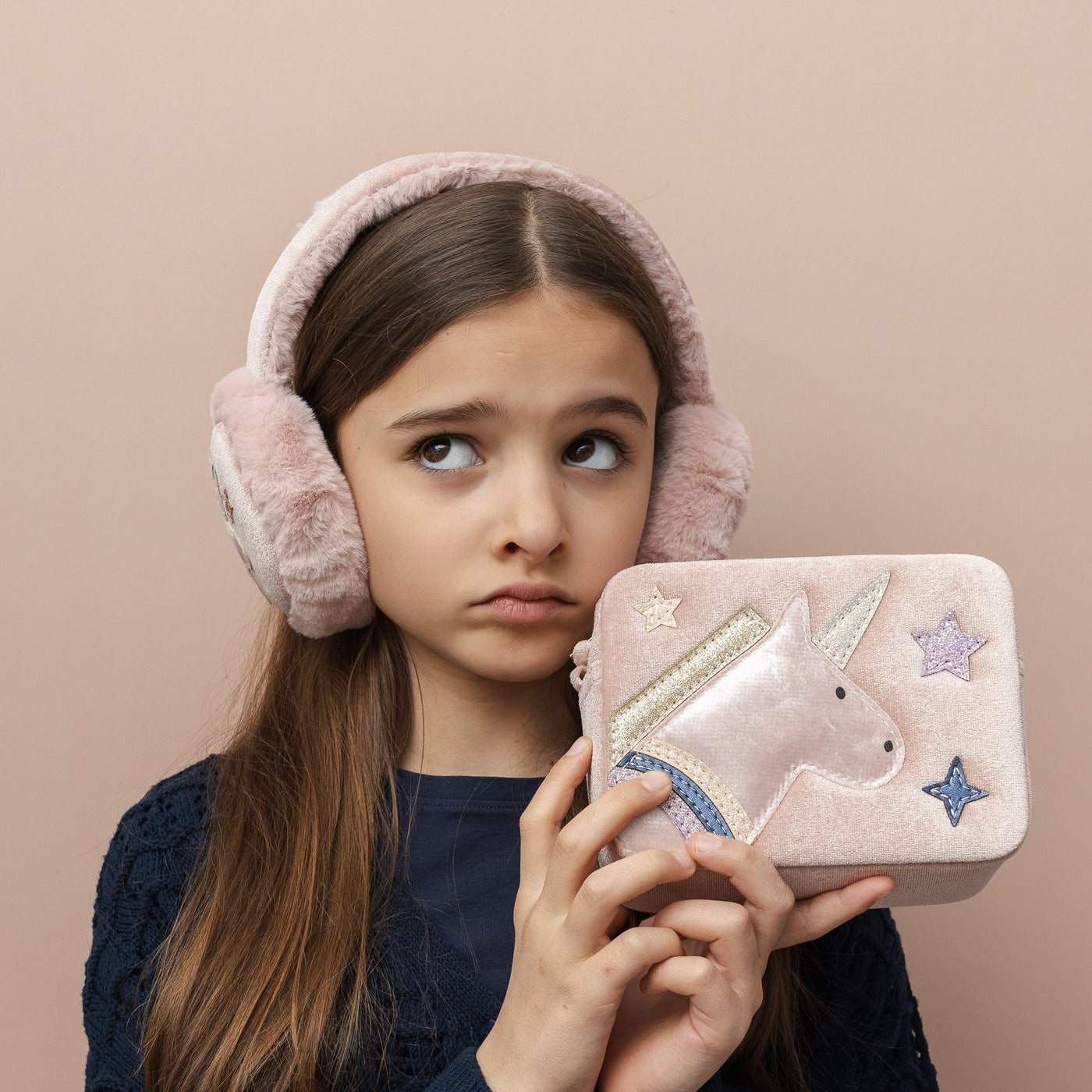 Girl wearing pink fluffy earmuffs holding a unicorn-themed bag against a beige background