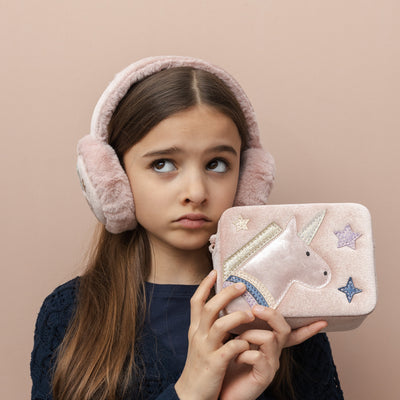 Girl wearing pink fluffy earmuffs holding a unicorn-themed bag against a beige background