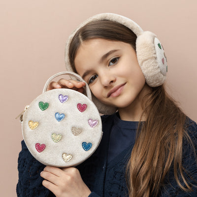 Dark haired girl holding a soft velvet, neutral coloured round bag embellished with multicoloured glitter and metallic love hearts with carry handle and cross body strap, also wearing fluffy cream matching earmuffs with jazzy heart print design