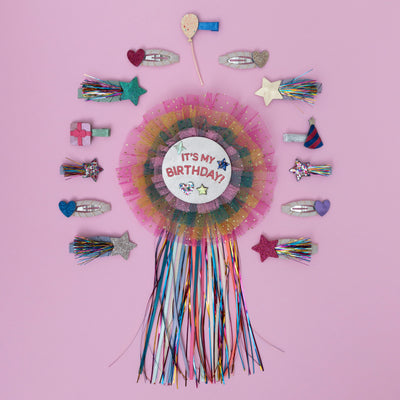 A vibrant rainbow birthday badge, bursting with a spectrum of bold colours, surrounded by a fun assortment of party-themed hair clips, perfect for adding a cheerful touch to any celebration