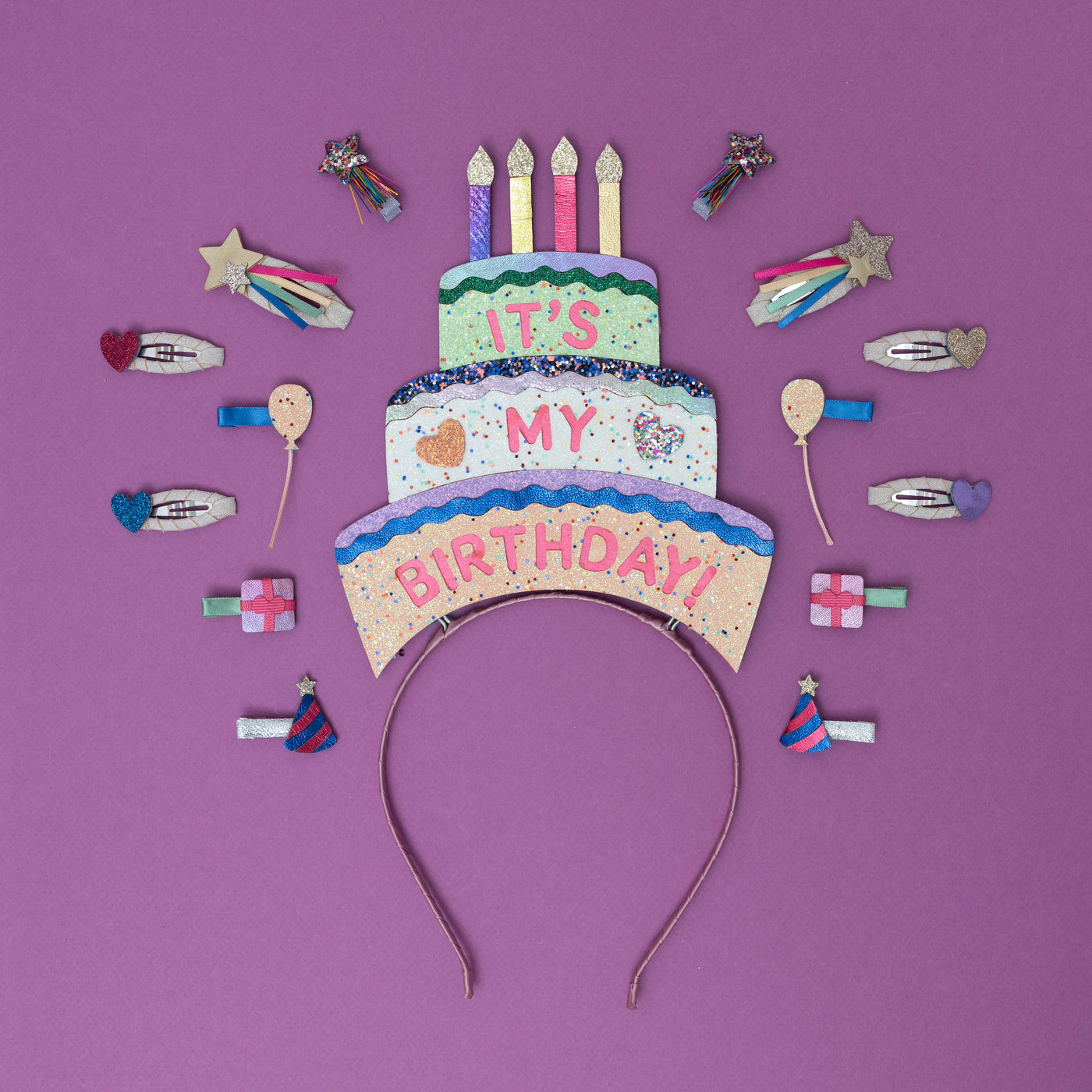 A sparkling, three-tiered glittery birthday cake headpiece, adorned with colourful candles and shimmering details, surrounded by festive party-themed hair clips
