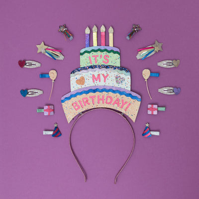 A sparkling, three-tiered glittery birthday cake headpiece, adorned with colourful candles and shimmering details, surrounded by festive party-themed hair clips