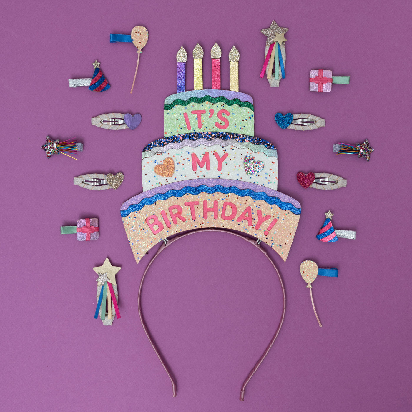 A sparkling, three-tiered glittery birthday cake headpiece, adorned with colourful candles and shimmering details, surrounded by festive party-themed hair clips