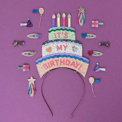 A sparkling, three-tiered glittery birthday cake headpiece, adorned with colourful candles and shimmering details, surrounded by festive party-themed hair clips