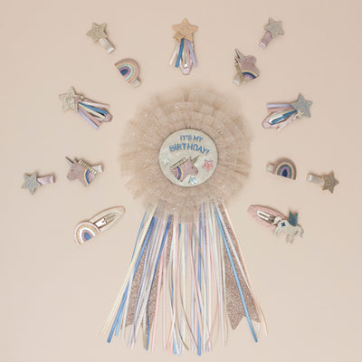 A magical unicorn birthday badge, featuring a sparkling unicorn design and encircled by glitter-speckled pink tulle, surrounded by an array of unicorn-themed hair clips, featuring glittery horns and manes, and whimsical stars, perfect for adding a fantastical touch to any celebration