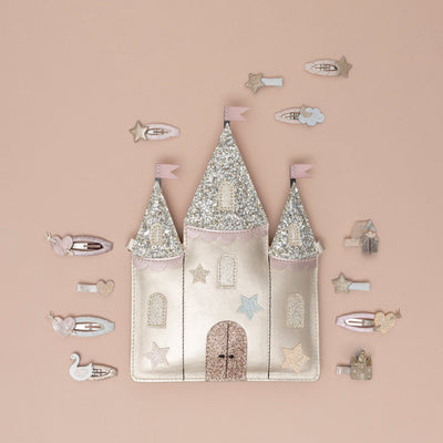 An enchanted gold castle bag, featuring glittery spires with pink flags and a cream cord crossbody strap, surrounded by a collection of princess and castle-themed hair clips