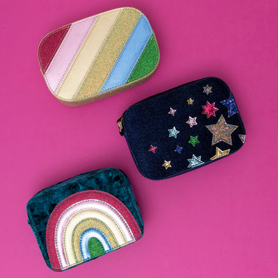 A trio of vibrant bags: a dazzle rainbow stripe bag, a navy velvet night sky bag, and a radiant blue rainbow velvet bag, each showcasing a bold mix of colours and textures, set against a bright pink background