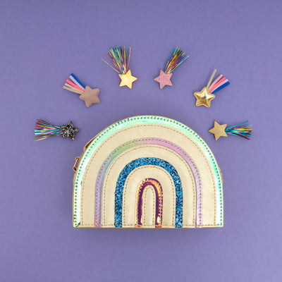 Brightly coloured gold metallic rainbow shaped children's handbag on a purple background with ribbon and tinsel shooting star hair clips place above