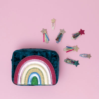 A radiant teal rainbow velvet bag, encircled by sparkling glitter star-themed hair clips, all set against a soft pink background, creating a dreamy and whimsical display