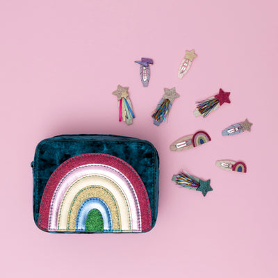 A radiant teal rainbow velvet bag, encircled by sparkling glitter star and rainbow themed hair clips, all set against a soft pink background, creating a dreamy and whimsical display