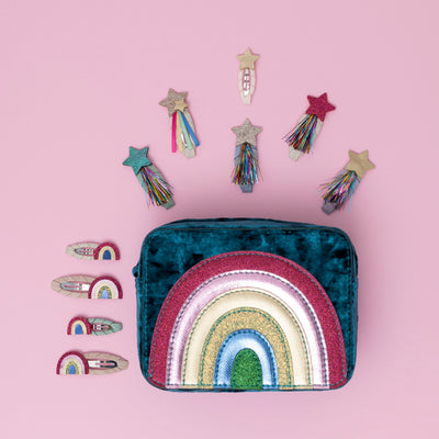 A radiant teal rainbow velvet bag, encircled by sparkling glitter star and rainbow themed hair clips, all set against a soft pink background, creating a dreamy and whimsical display
