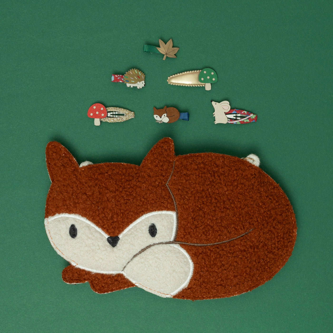 A Felix fox crossbody bag, surrounded by a delightful collection of woodland-themed hair accessories, including a hedgehog, toadstool, fox, mouse, and autumn leaf, all set against a rich moss green background