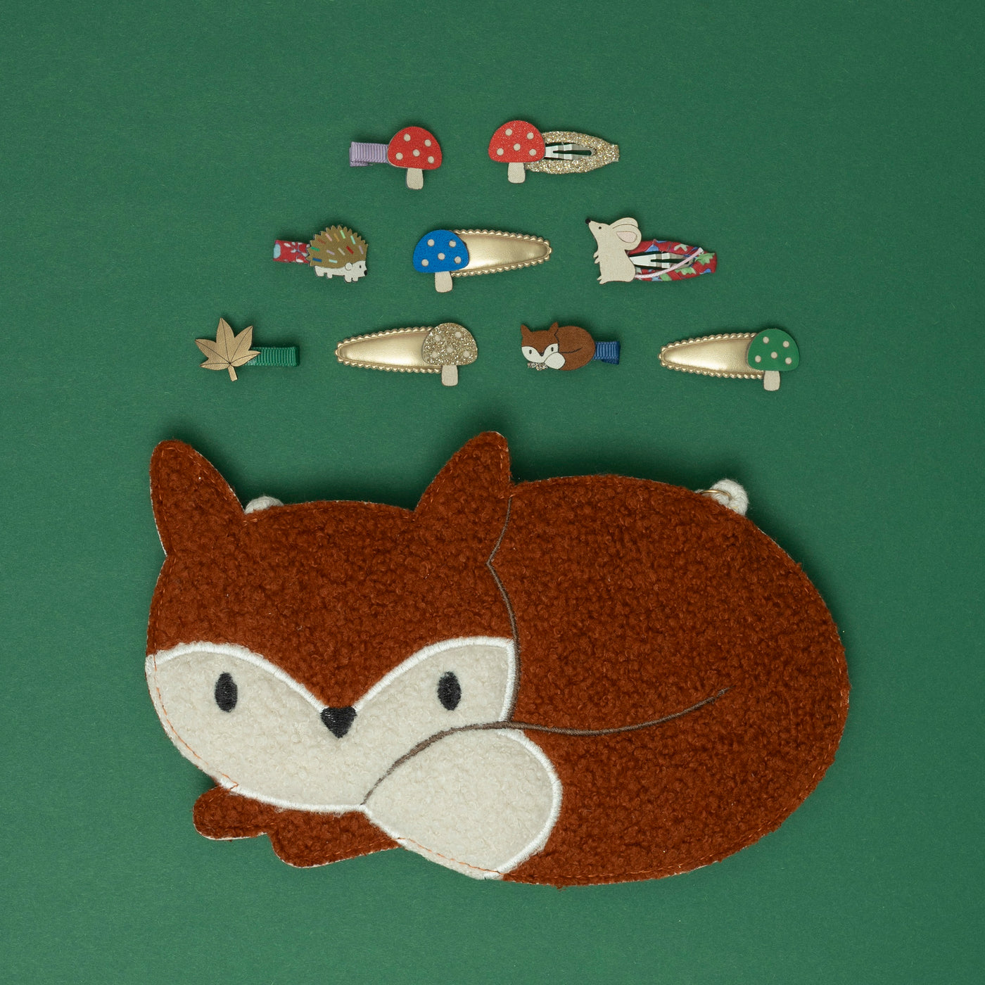 A Felix fox crossbody bag, surrounded by a delightful collection of woodland-themed hair accessories, including a hedgehog, toadstool, fox, mouse, and autumn leaf, all set against a rich moss green background