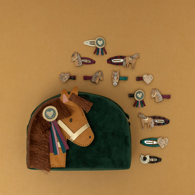 A green velvet pony pal bag with a matching green velvet strap, surrounded by an assortment of pony-themed hair clips, set against a warm golden yellow background