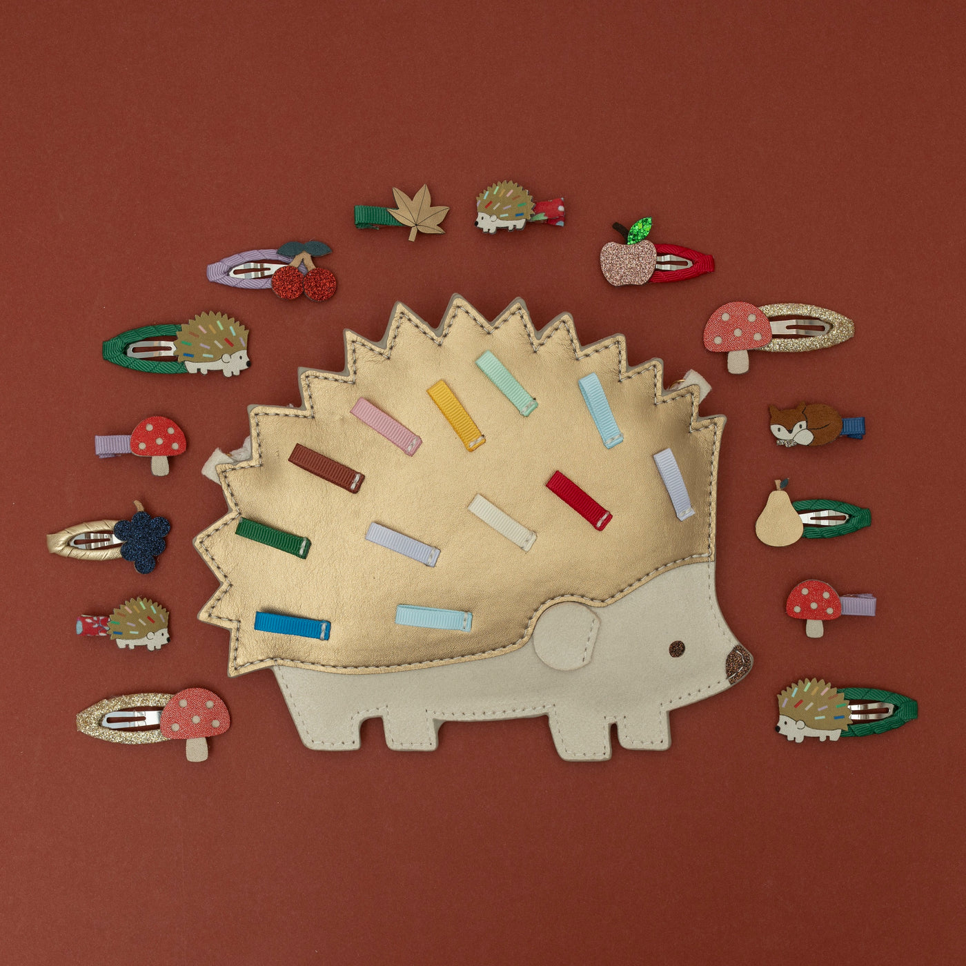 A Harry hedgehog bag in gold and cream leatherette, surrounded by a collection of woodland-themed and orchard fruit-themed hair clips, including a hedgehog, toadstool, fox, autumn leaf, and various fruit designs, all set against a rich chestnut brown background