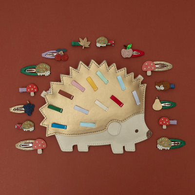 A Harry hedgehog bag in gold and cream leatherette, surrounded by a collection of woodland-themed and orchard fruit-themed hair clips, including a hedgehog, toadstool, fox, autumn leaf, and various fruit designs, all set against a rich chestnut brown background