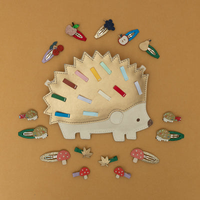 A Harry hedgehog bag in gold and cream leatherette, surrounded by a collection of woodland-themed and orchard fruit-themed hair clips, including a hedgehog, toadstool, fox, autumn leaf, and various fruit designs, all set against a warm golden yellow background