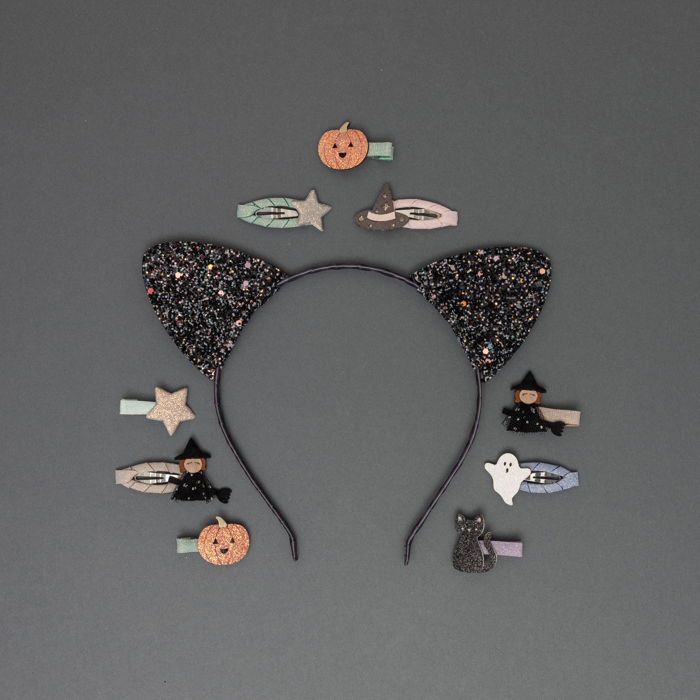 Glittery black cat ears on a sateen ribbon-covered headband, surrounded by Halloween-themed hair clips, including a witch on a broomstick, witch hat, pumpkin, ghost, black cat, and stars, all crafted from glittery fabrics, set against a soft dark grey background