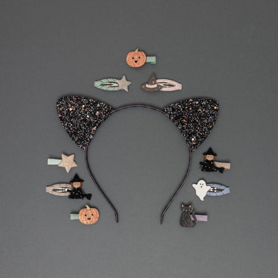 Glittery black cat ears on a sateen ribbon-covered headband, surrounded by Halloween-themed hair clips, including a witch on a broomstick, witch hat, pumpkin, ghost, black cat, and stars, all crafted from glittery fabrics, set against a soft dark grey background