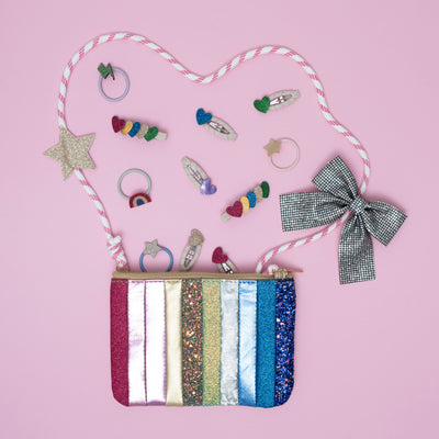 Stripe bag in glitter and metallic fabrics with bungee cord cross body strap on pink background surrounded by disco ball effect bow hair clip and smaller layered glitter heart clips and single heart clips and ponies