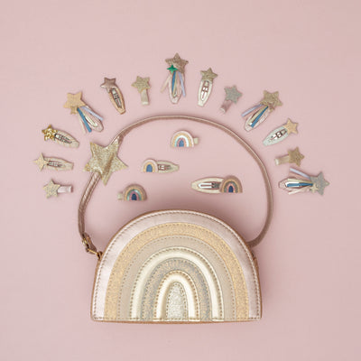 A glittery pink rainbow bag surrounded by a sparkling collection of magical rainbow, and star hair clips, set against a pale pink background