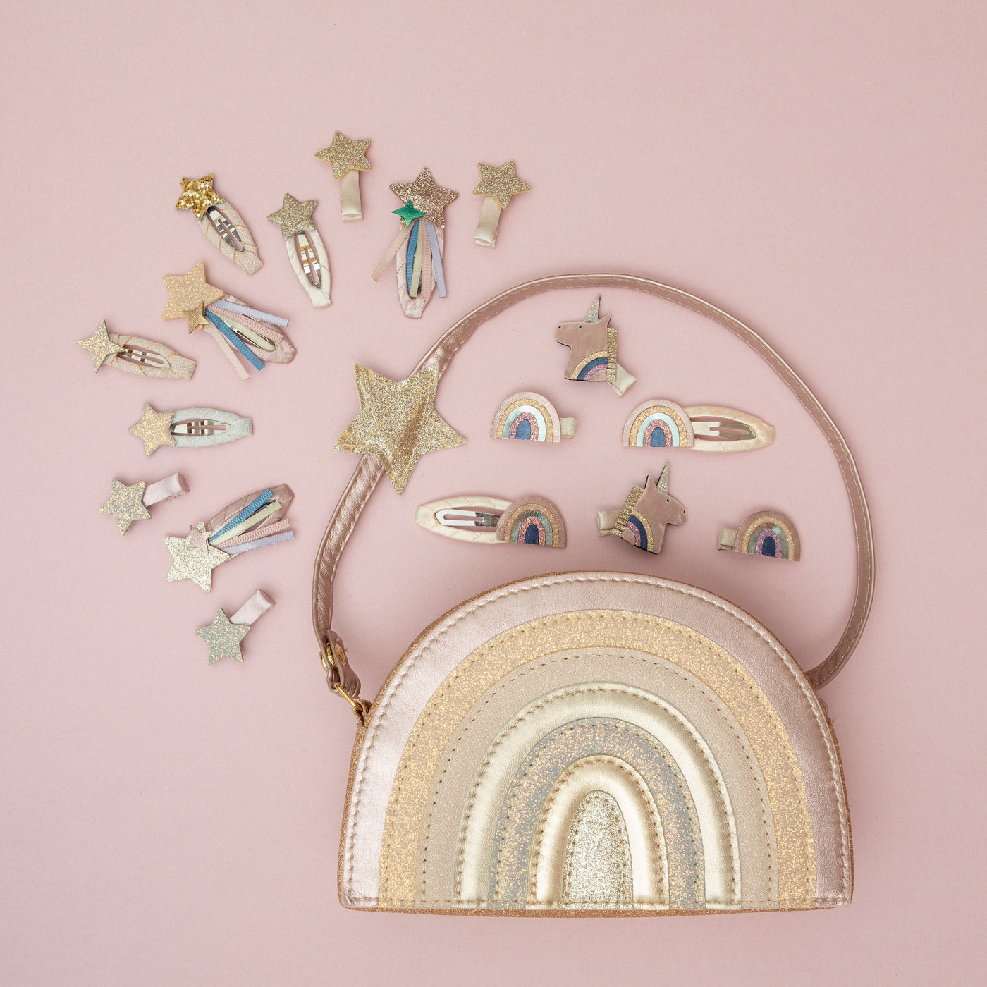 A glittery pink rainbow bag surrounded by a sparkling collection of magical unicorn, rainbow, and star hair clips, set against a pale pink background