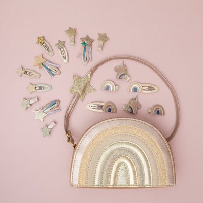 A glittery pink rainbow bag surrounded by a sparkling collection of magical unicorn, rainbow, and star hair clips, set against a pale pink background