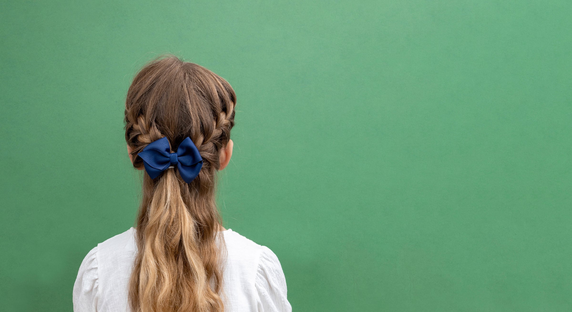 A child viewed from behind wearing a white top and a long blonde ponytail secured with a dark blue bow, standing against a solid green background.
