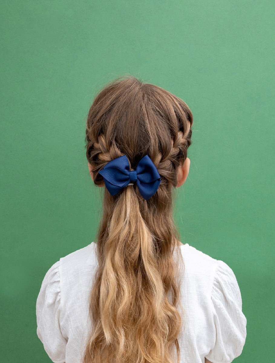 A child viewed from behind wearing a white blouse, with long wavy light brown hair styled in braids and a large dark blue bow accessory at the back of the head, set against a solid green background.