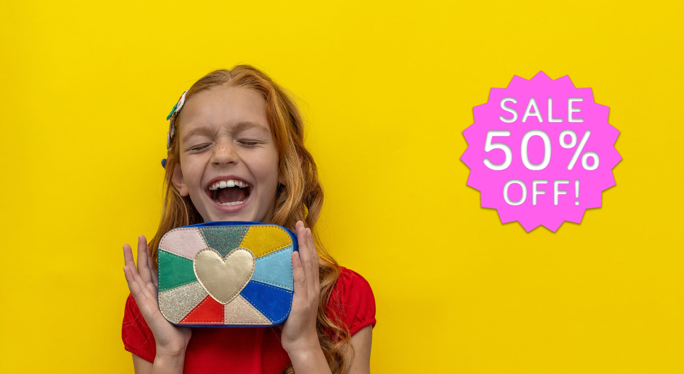A young girl with red hair is smiling and holding a colorful, patchwork-style heart-shaped purse against a bright yellow background, with a pink 'SALE 50% OFF!' badge on the right.