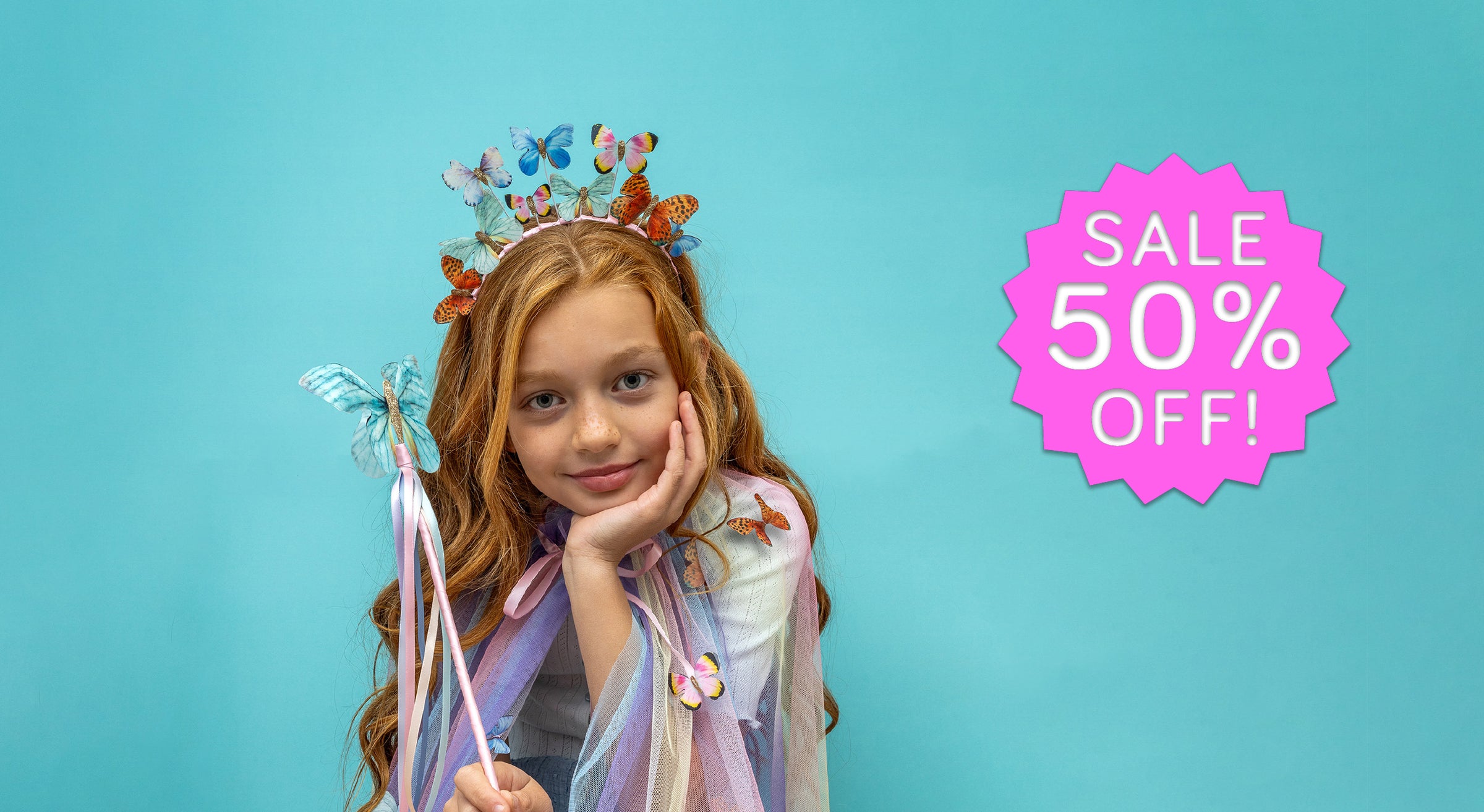 A young girl with long light brown hair poses in a whimsical fairy costume against a solid turquoise background. She is wearing a butterfly crown and holding a butterfly wand.