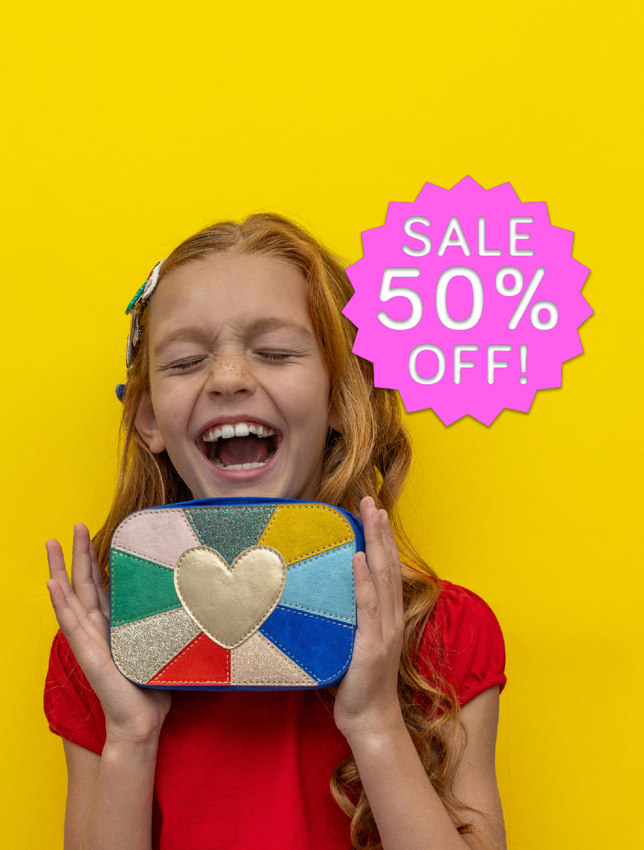 A young girl with red hair holds up a colorful patchwork clutch purse with a glittery gold heart in the center. She is wearing a red shirt against a bright yellow background, and a pink starburst graphic on the right reads 'SALE 50% OFF!'.
