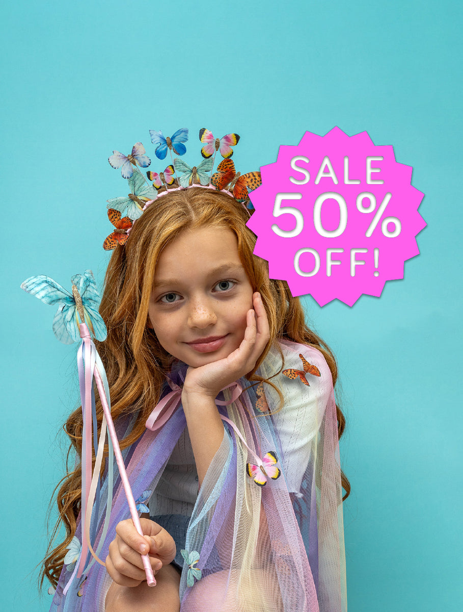 A young girl with long light brown hair poses in a whimsical fairy costume against a solid turquoise background. She is wearing a butterfly crown, a sheer lavender and blue cape, and holding a butterfly-tipped wand.