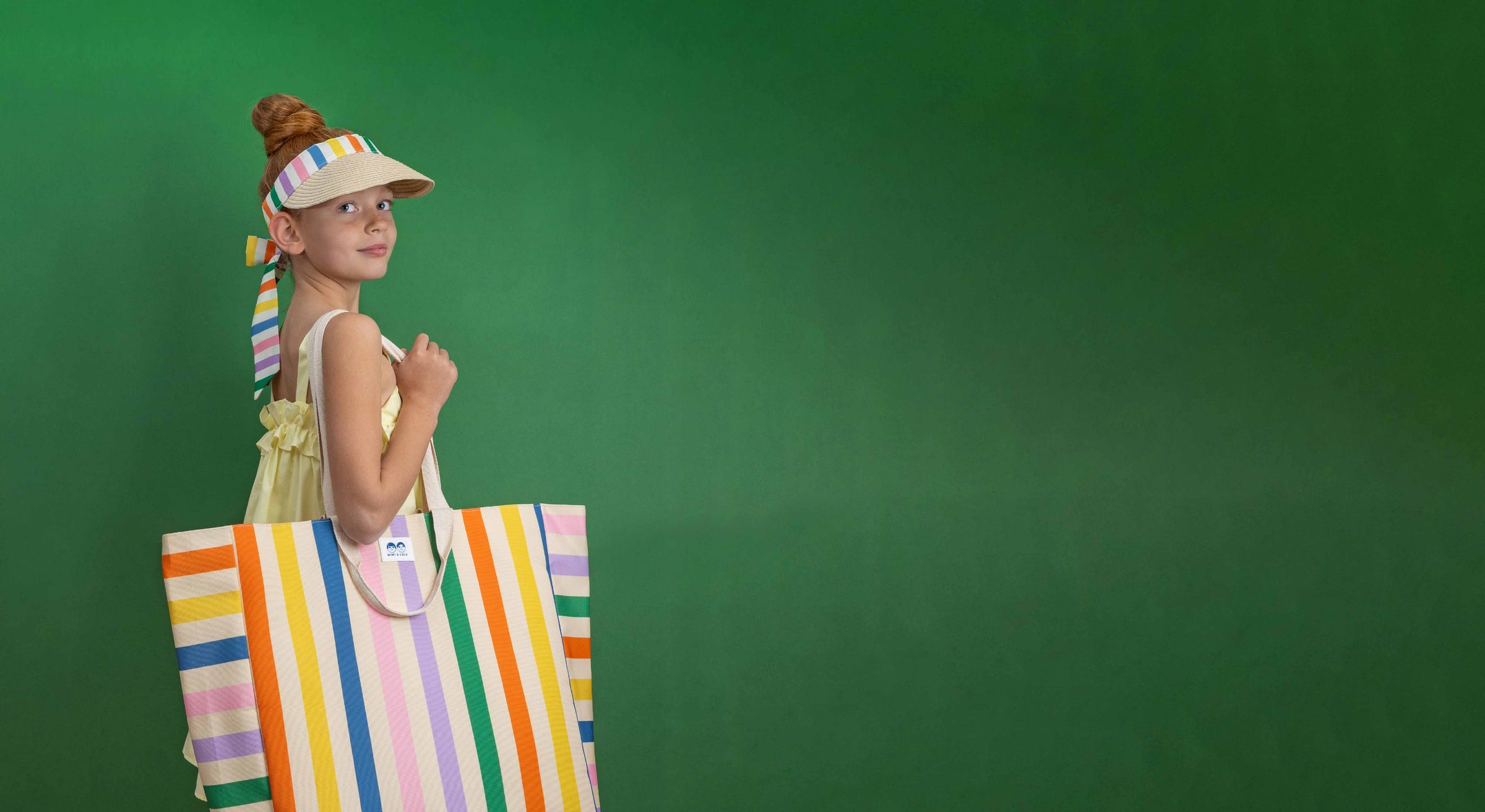 A girl wearing Mimi & Lula's stripe visor and strip beach bag against a green background