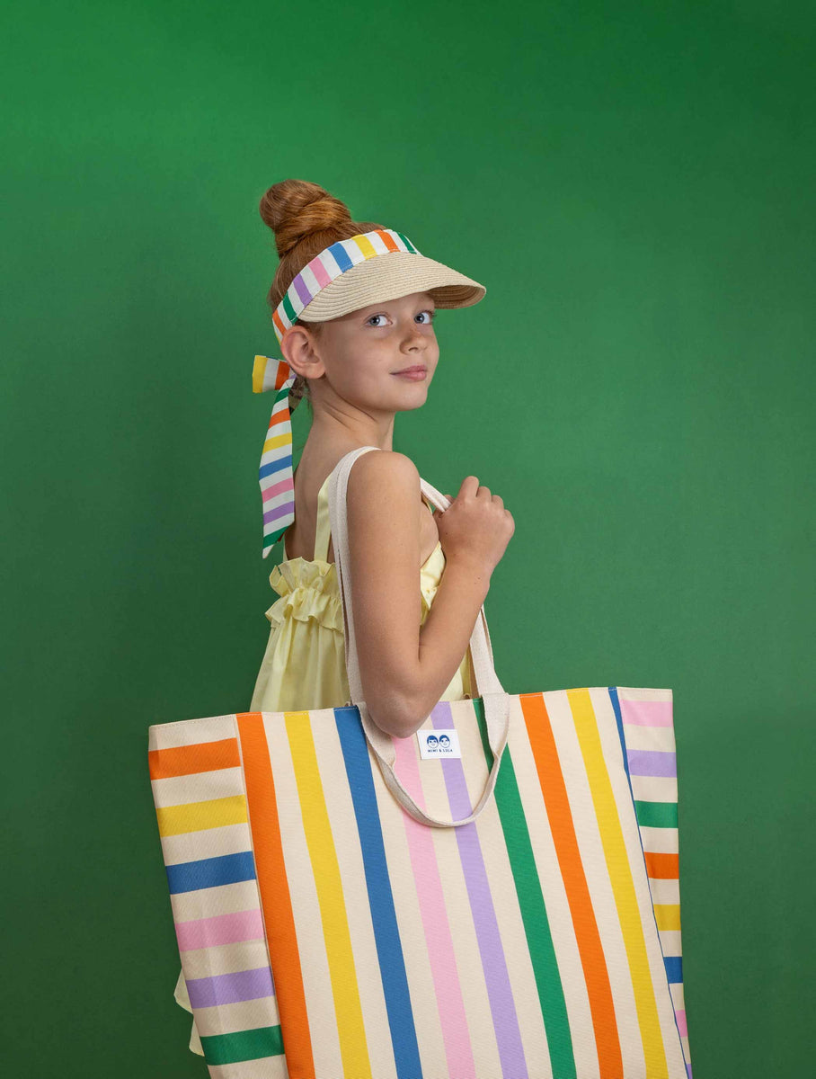 Girl wearing Mimi & Lula's stripe visor and strip beach bag against a green background