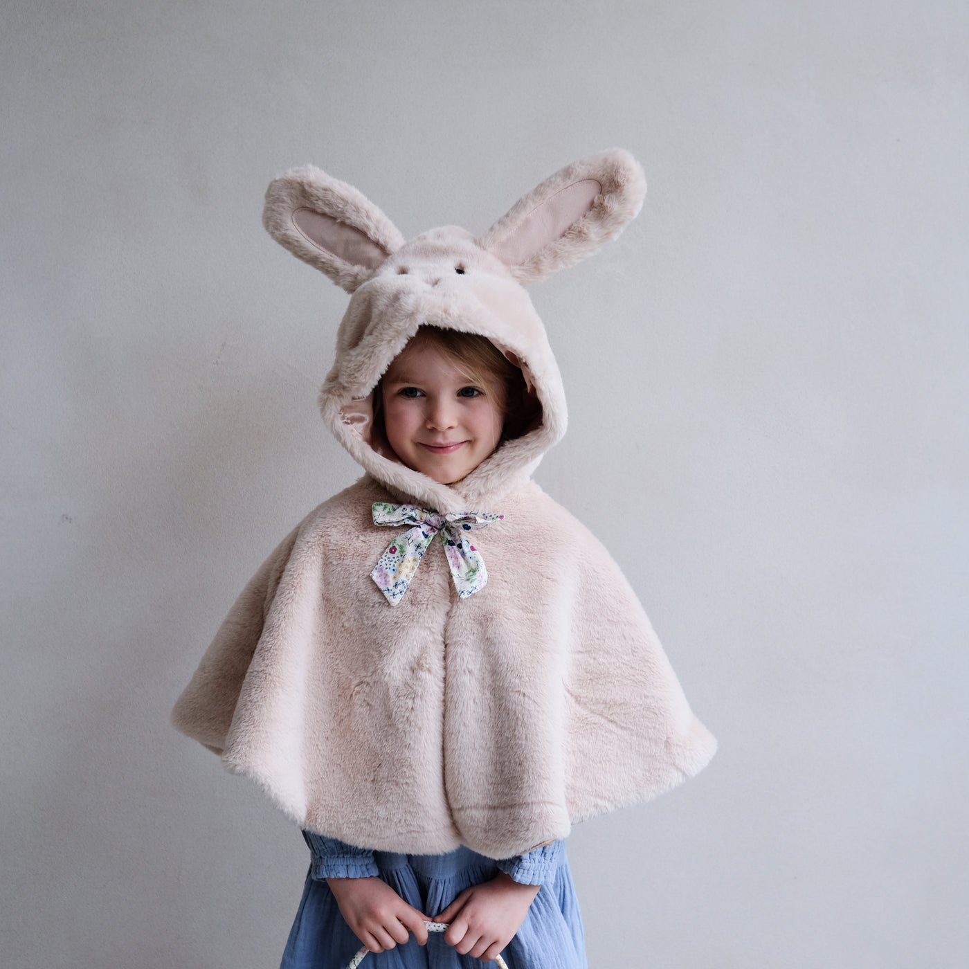 Smiling little girl wearing a fluffy bunny cape for dressing up with hood including embroidered face, fluffy ears and a floral print fabric bow with popper to neck and satin lining