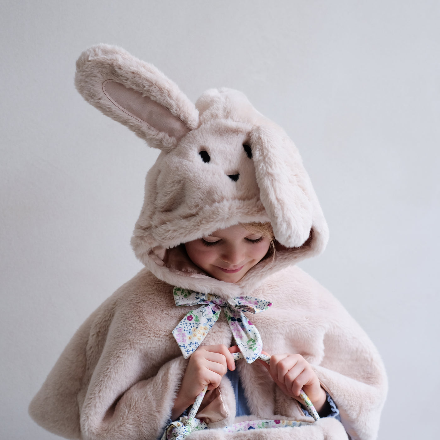 Little girl looking into fluffy bunny basket wearing a fluffy bunny cape for dressing up with hood including embroidered face, fluffy ears and a floral print fabric bow with popper to neck and satin lining