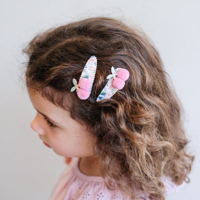 Little girl wearing a pair of floral print hair clips featuring a pair of soft pink pom pom cherries to the side