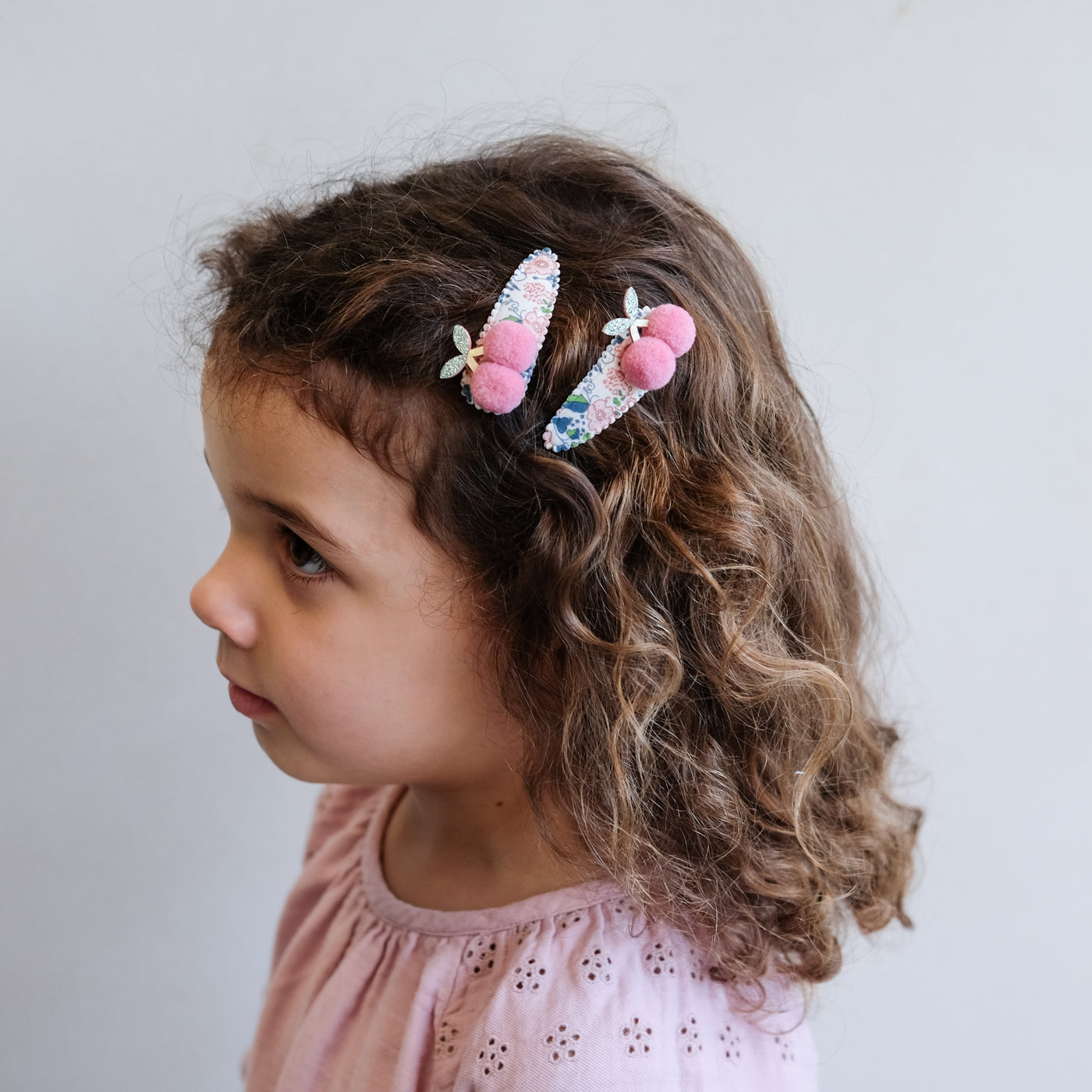 Little girl wearing a pair of floral print hair clips featuring a pair of soft pink pom pom cherries to the side