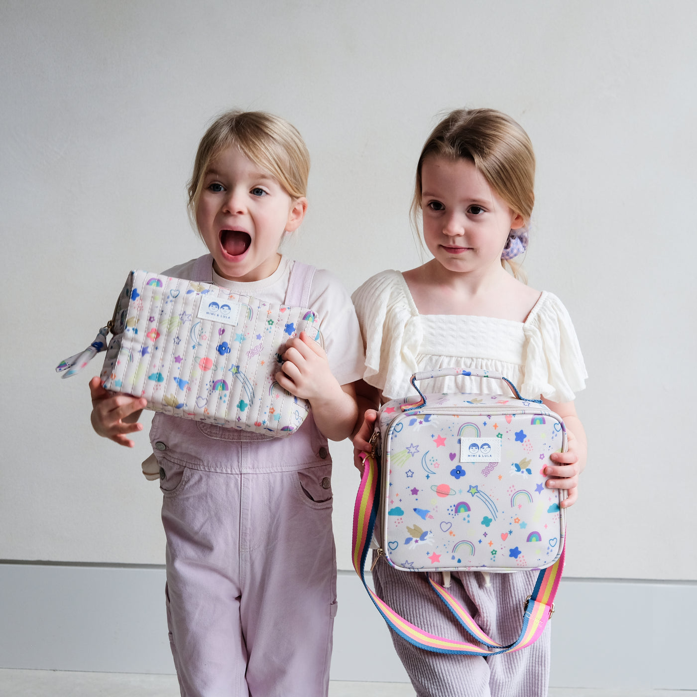 Two little girls holding a large wash bag with zip and lunch bag with rainbow strap both in a bright and fun unicorn and rainbow print fabric 