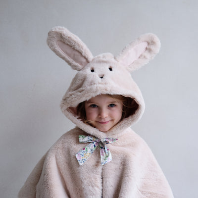 Smiling little girl wearing a cute fluffy bunny cape with hood up showing two fluffy bunny ears and embroidered face with floral bow to the neck